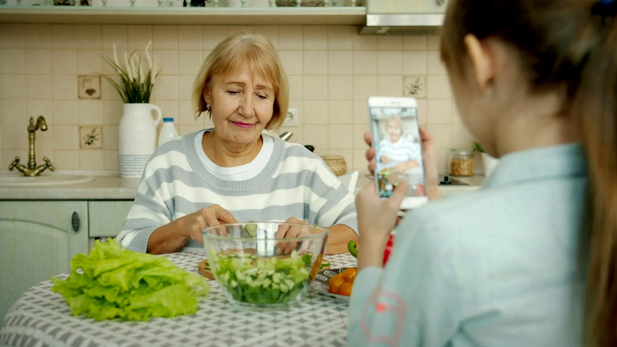A granddaughter films her grandmother on a phone while she makes a salad at the kitchen table