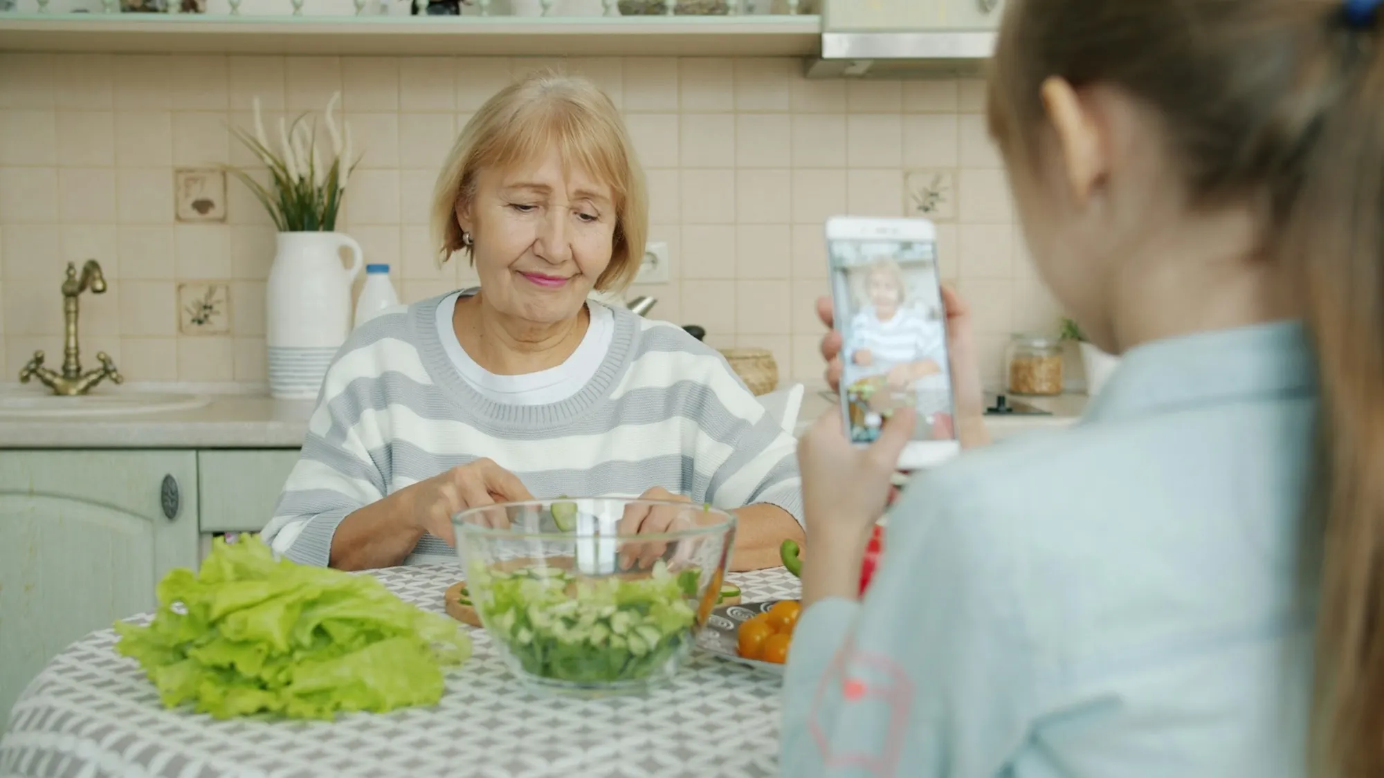 A granddaughter films her grandmother on a phone while she makes a salad at the kitchen table