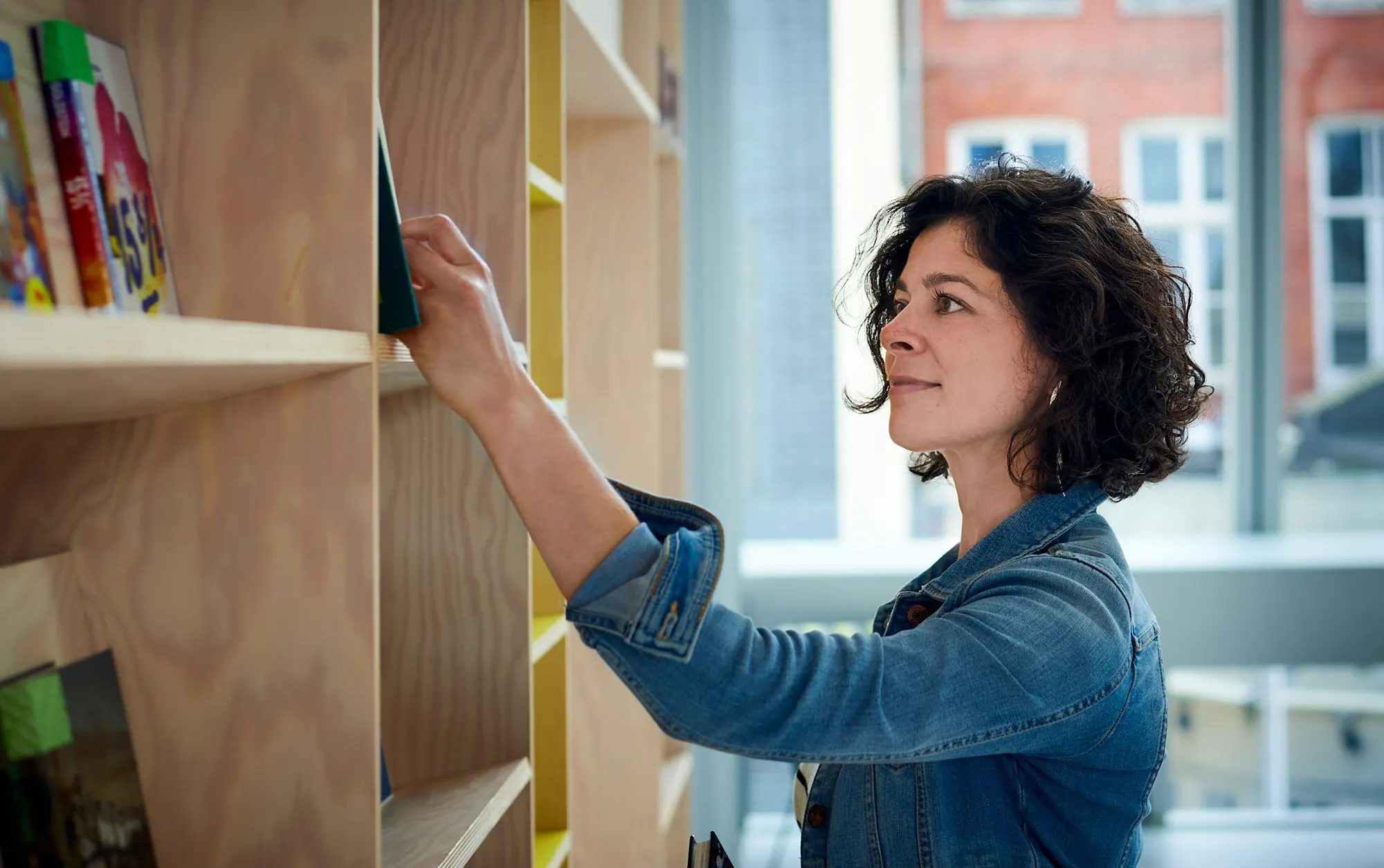 A woman pulls a book from a light wood bookshelf by a sunny window