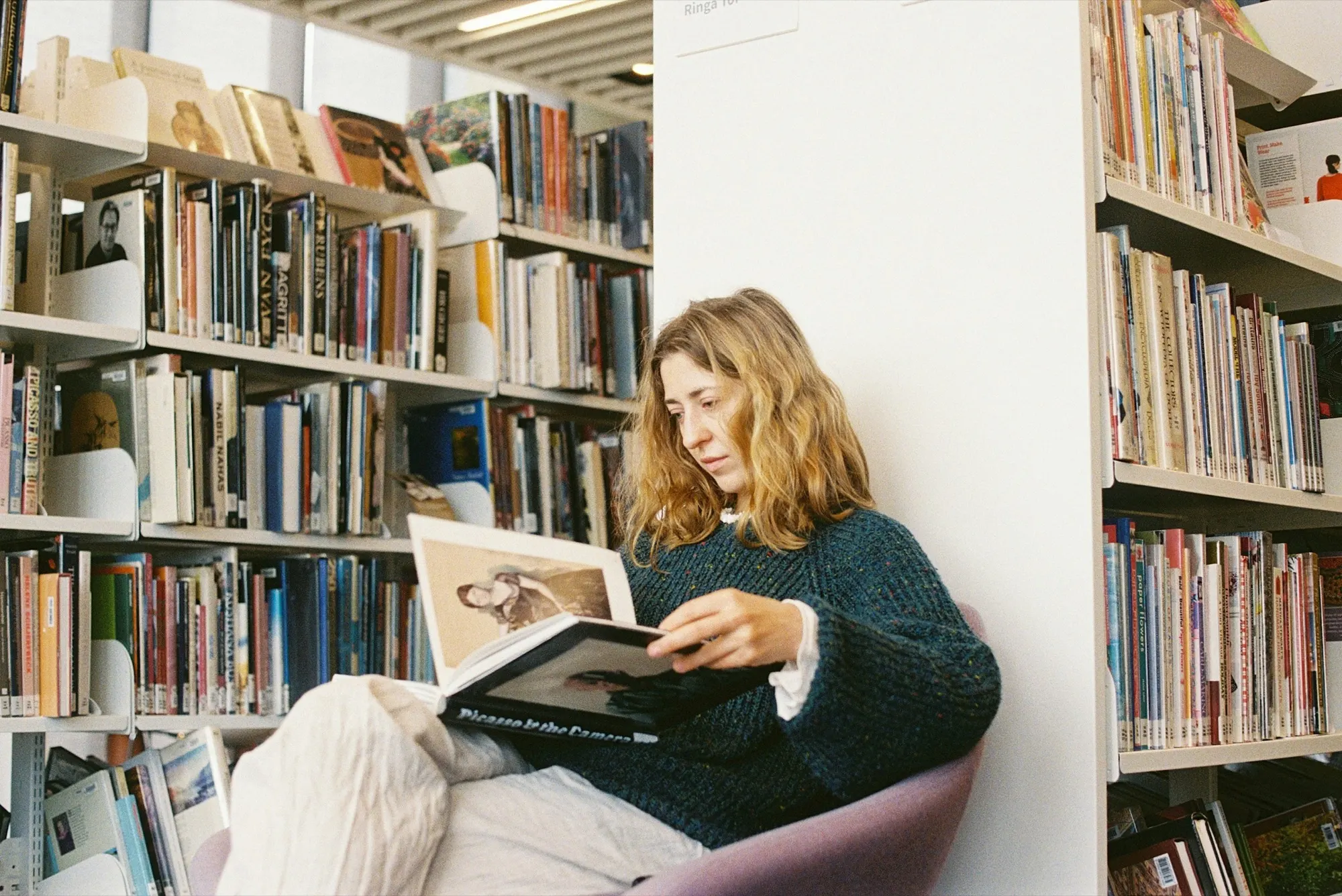 A woman reading a photo book, curled up between tall shelves of books